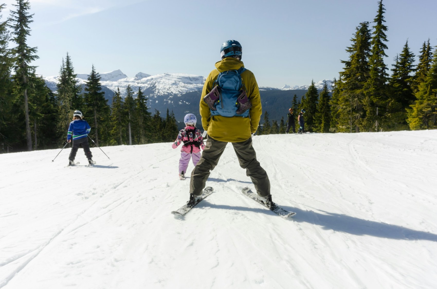 An adult in a yellow jacket and two children skiing a wide groomed run, with evergreens and distant snowy peaks