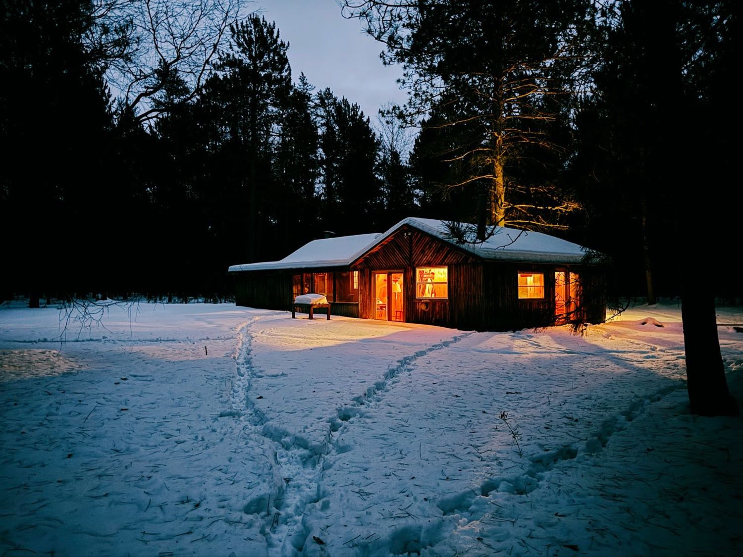 A low timber building with warmly lit windows sitting in deep snow among tall pines at dusk