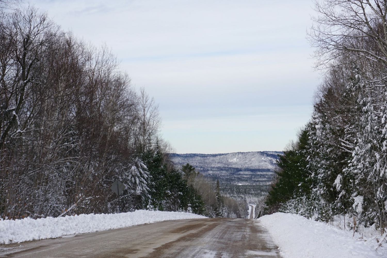 A snow-banked two-lane road descending through winter forest towards low forested hills